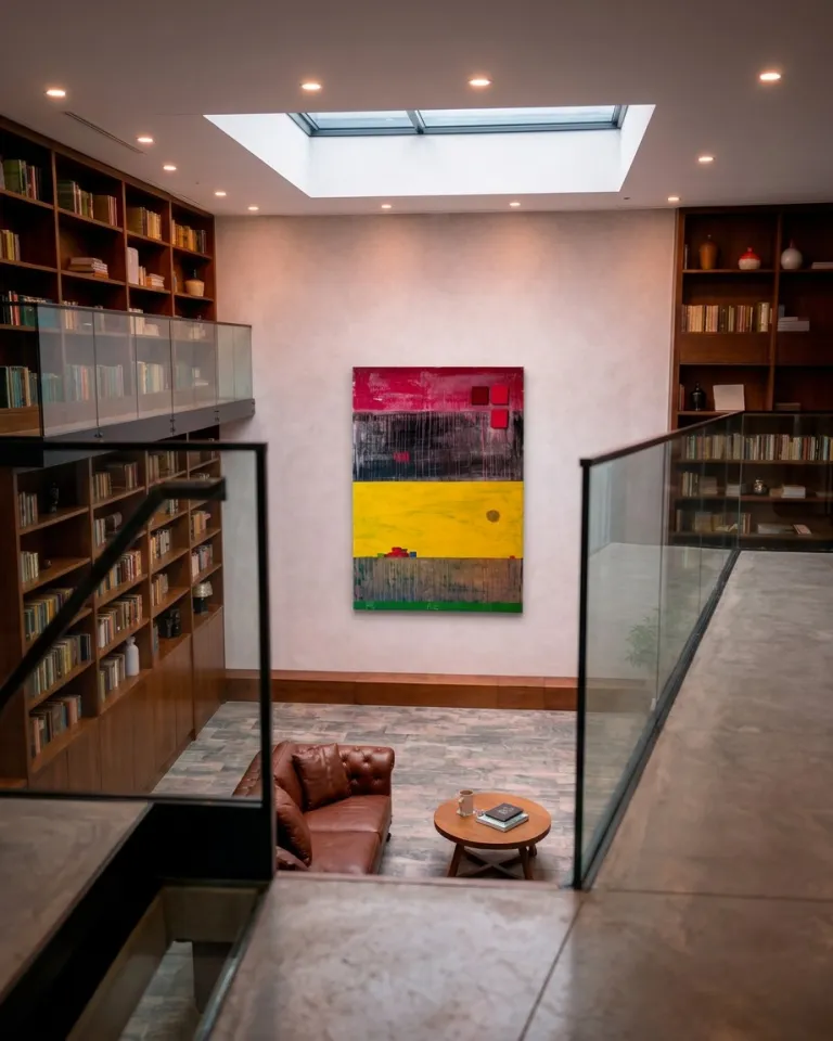 Vertical abstract painting by Maurizio Valch featuring horizontal bands of red, black, and yellow, with brown and green details. Displayed on a light wall in a modern, double-height library, alongside wooden bookshelves, a glass mezzanine railing, and a central skylight. A leather sofa and a wooden table are visible below.