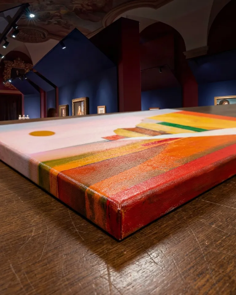Close table-level view of VIA SOLIS by Maurizio Valch, with the canvas edge visible, a white path, orange and yellow blocks, and an orange sun against a pink ground.