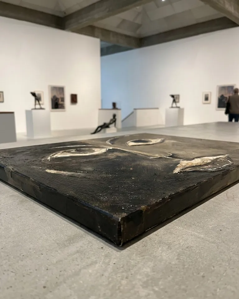 Raking perspective of the canvas lying on a gray slab in a museum room, highlighting the relief of the incised marks of the eyes and mouth.