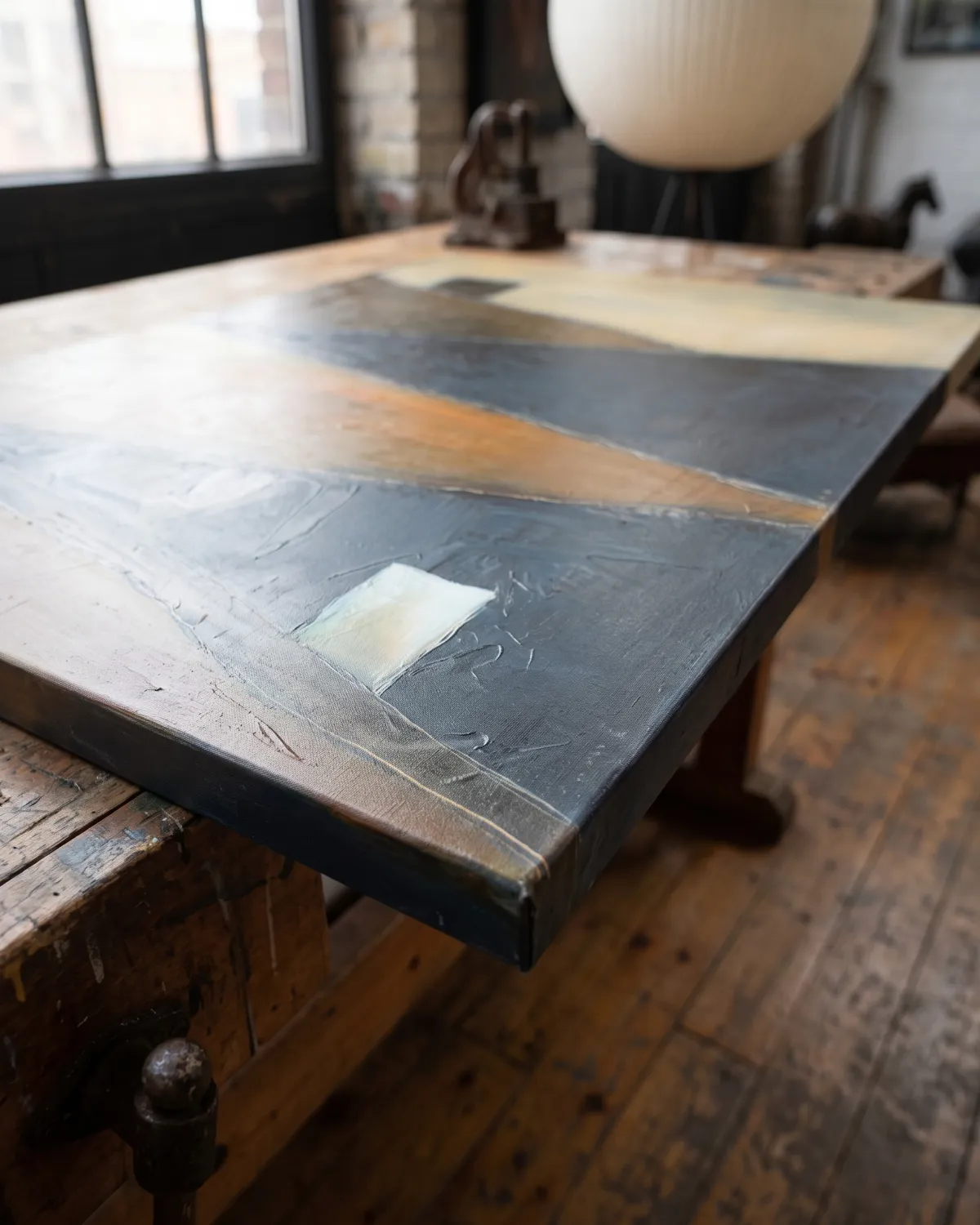 Vista en perspectiva oblicua y a ras de suelo de una pintura abstracta apoyada sobre un banco de trabajo de madera en un taller industrial. En primer plano, destaca la esquina inferior derecha del lienzo con un pequeño rectángulo blanco luminoso en relieve y el canto lateral pintado en color oscuro. La superficie del lienzo, con texturas de acrílico y óleo en tonos azul y ocre, se extiende en diagonal hacia el fondo en un suave desenfoque. Al fondo, se aprecian un gran ventanal y una lámpara esférica.