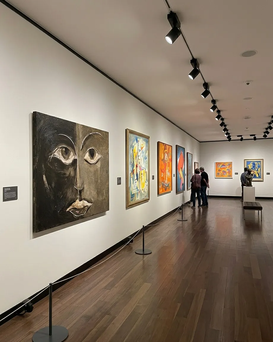 Perspectiva de la obra de Maurizio Valch colgada en la pared blanca de un pasillo de museo iluminado con focos dirigidos y suelo de madera.