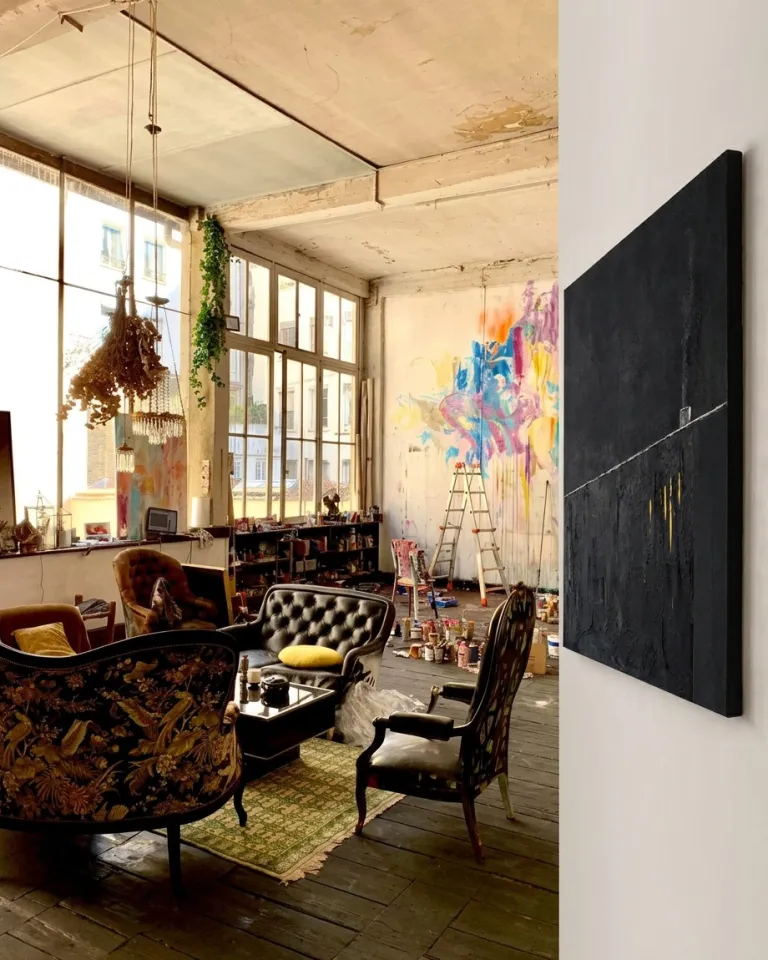Large-format black abstract painting hung in profile in a bright art workshop with antique black leather armchairs and a paint-splattered wall.