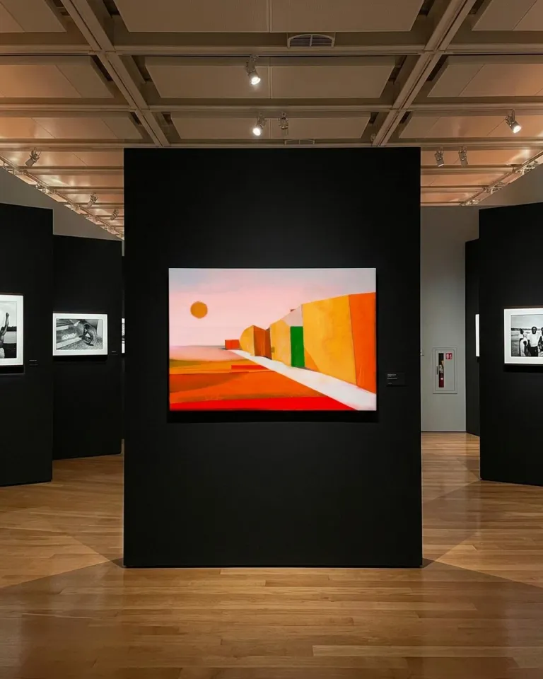 VIA SOLIS by Maurizio Valch displayed on a black museum panel; the painting shows a white path between red, yellow, and green planes beneath an orange sun.