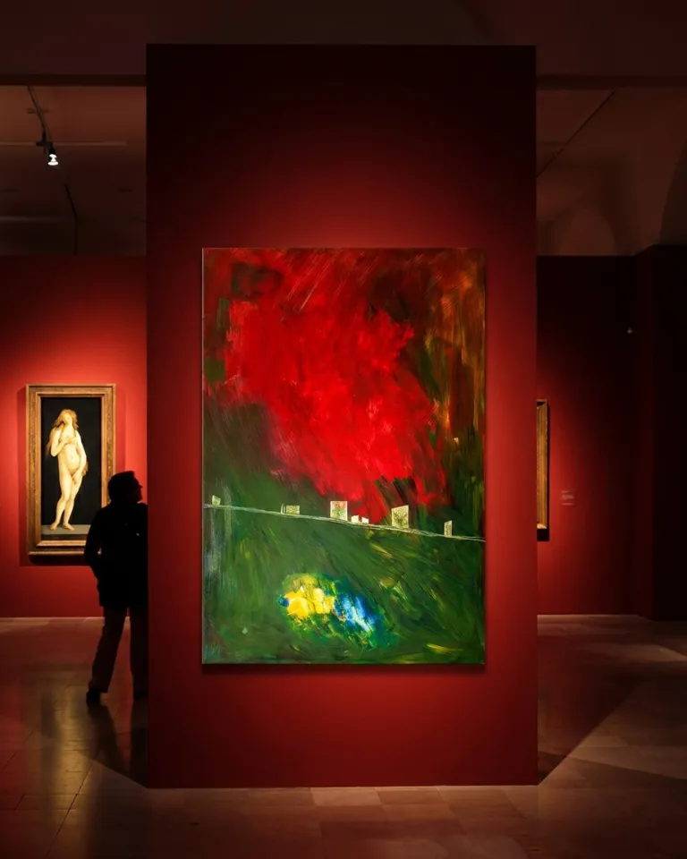 The artwork by Maurizio Valch displayed on a deep red interior wall in an art gallery, illuminated by focused spotlights with a person seen from behind contemplating it in the dim light.