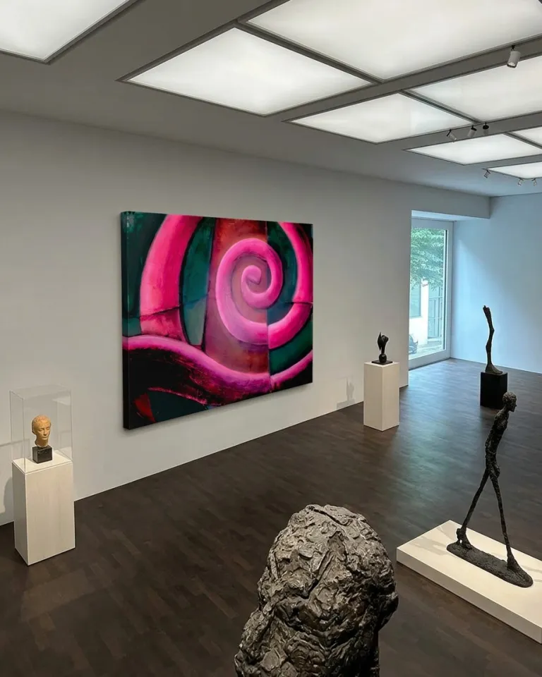 Vista aérea de la pintura abstracta VORAGO colgada en una galería de arte minimalista con esculturas exentas sobre pedestales, iluminación de techo y suelos de madera oscura.