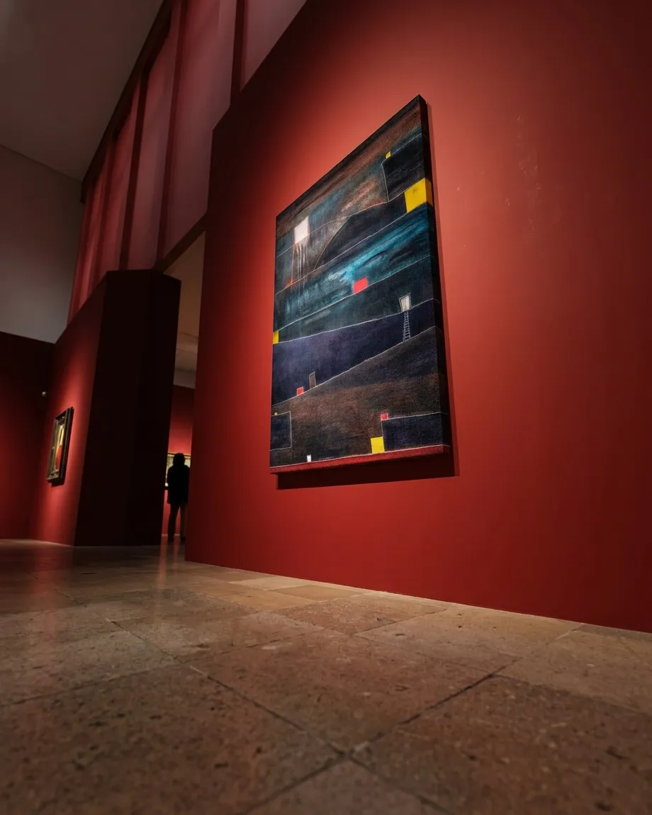 ADITUS de Maurizio Valch, pintura abstracta vertical de franjas oscuras, turquesa y pequeños acentos rojos y amarillos, instalada en la pared roja de un corredor de galería.
