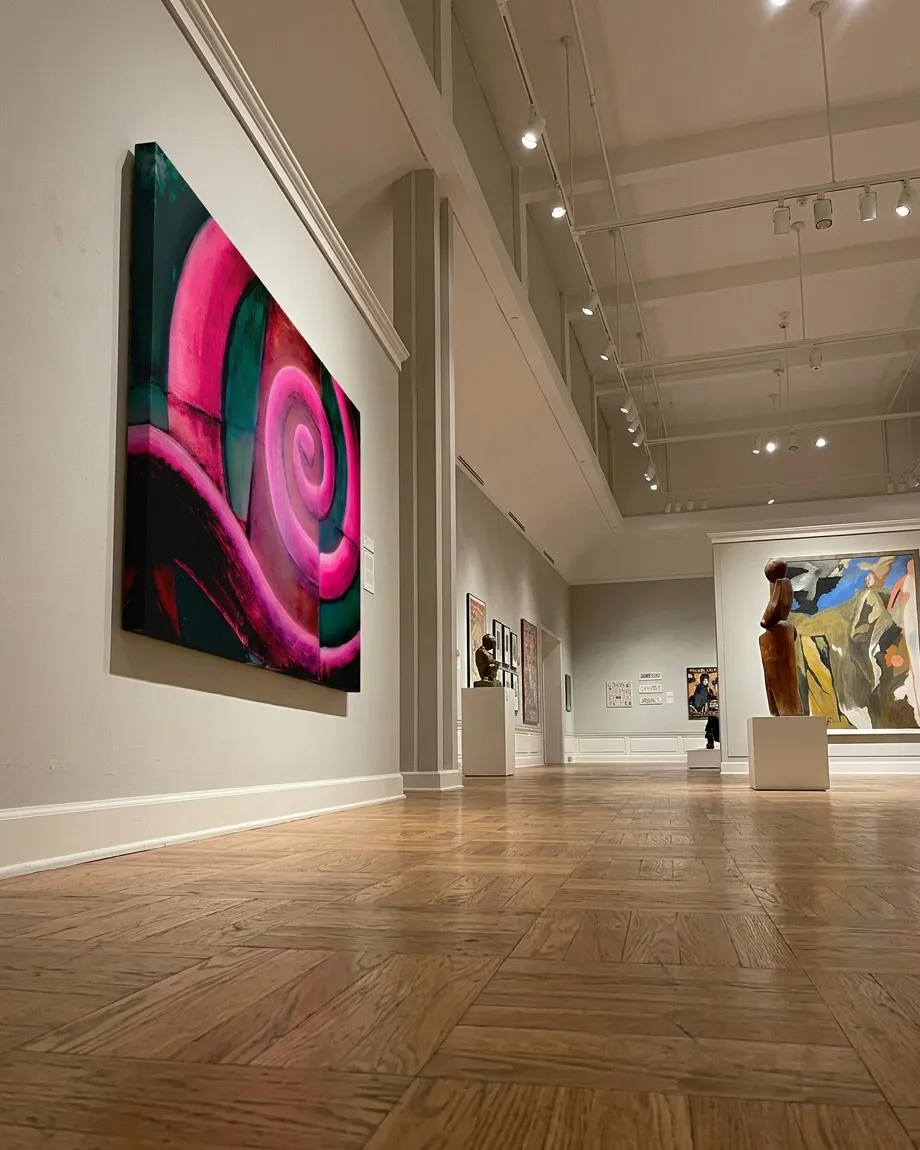 Perspectiva desde un ángulo bajo de la obra VORAGO montada en la pared blanca de una espaciosa galería de museo con suelos de parqué de madera y esculturas clásicas y modernas de fondo.