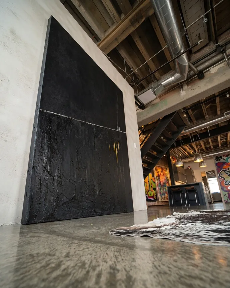 Large-format abstract painting leaning on the polished concrete floor of an industrial loft, next to a metal staircase and a ceiling with wooden beams.