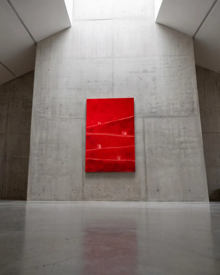 The red abstract painting ATHAR hanging on a monumental grey exposed concrete wall under a skylight in an industrial-style gallery.