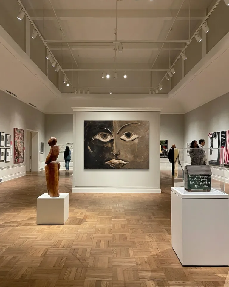A large abstract painting hanging on the central wall of an art gallery. It shows a face vertically divided into a black half and an ochre half, with almond-shaped eyes defined by clear incised lines.