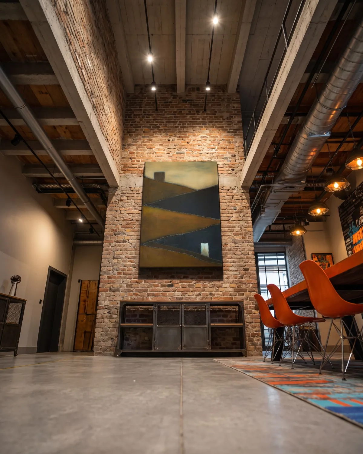 Vertical painting by Maurizio Valch hanging above an exposed brick fireplace in an industrial-style dining room with high wood and concrete ceilings.