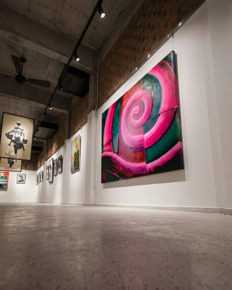 Fotografía en ángulo contrapicado de la pintura VORAGO expuesta en una galería de estilo industrial con vigas de hormigón, ladrillo visto, rieles de focos y suelo de cemento pulido.