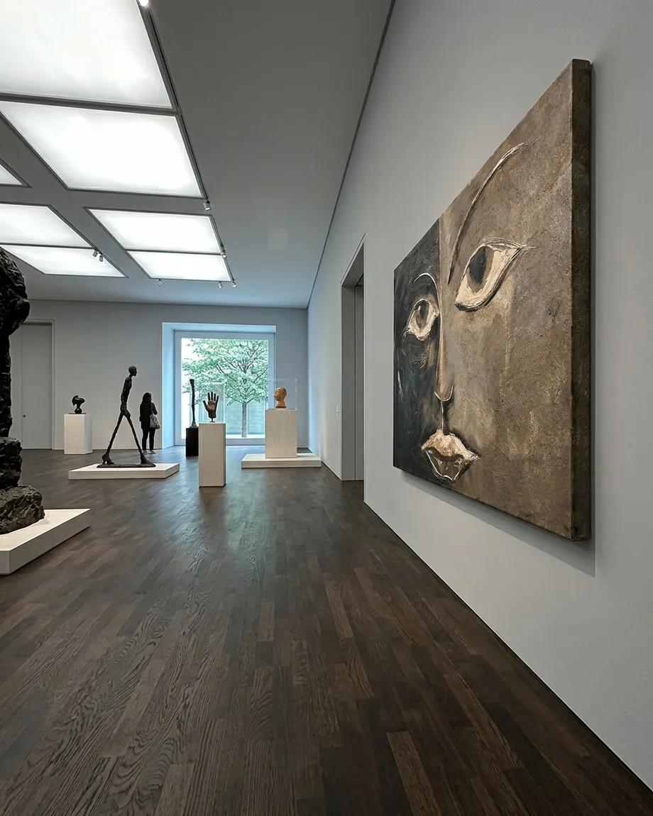 Cuadro de un rostro dividido en tonos ocre y negro expuesto oblicuamente en un museo minimalista con suelos de parquet oscuro y tragaluces.