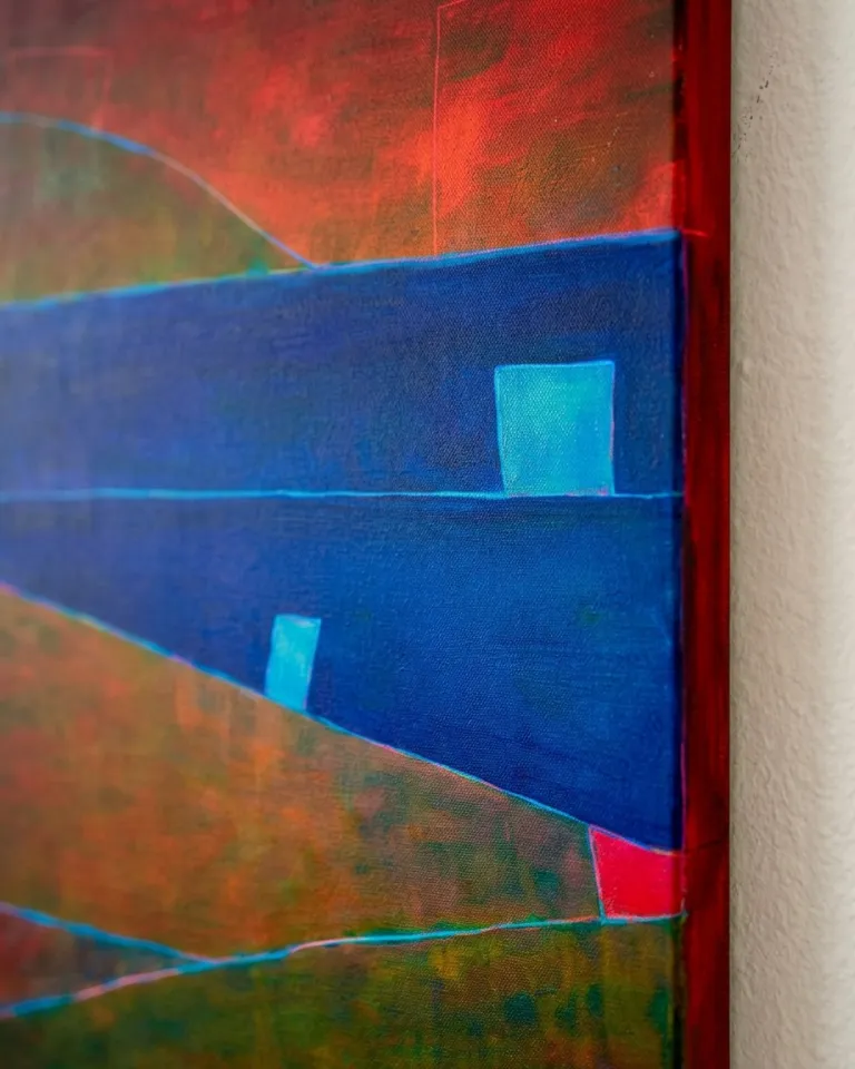 Fotografía de primer plano del canto pintado en rojo carmesí de un lienzo vertical. La toma lateral muestra el grosor del bastidor de tres centímetros junto a una pared de yeso texturizada, revelando campos de color azul cobalto, ocre, finas líneas incisas y formas celestes sobre la superficie del cuadro.