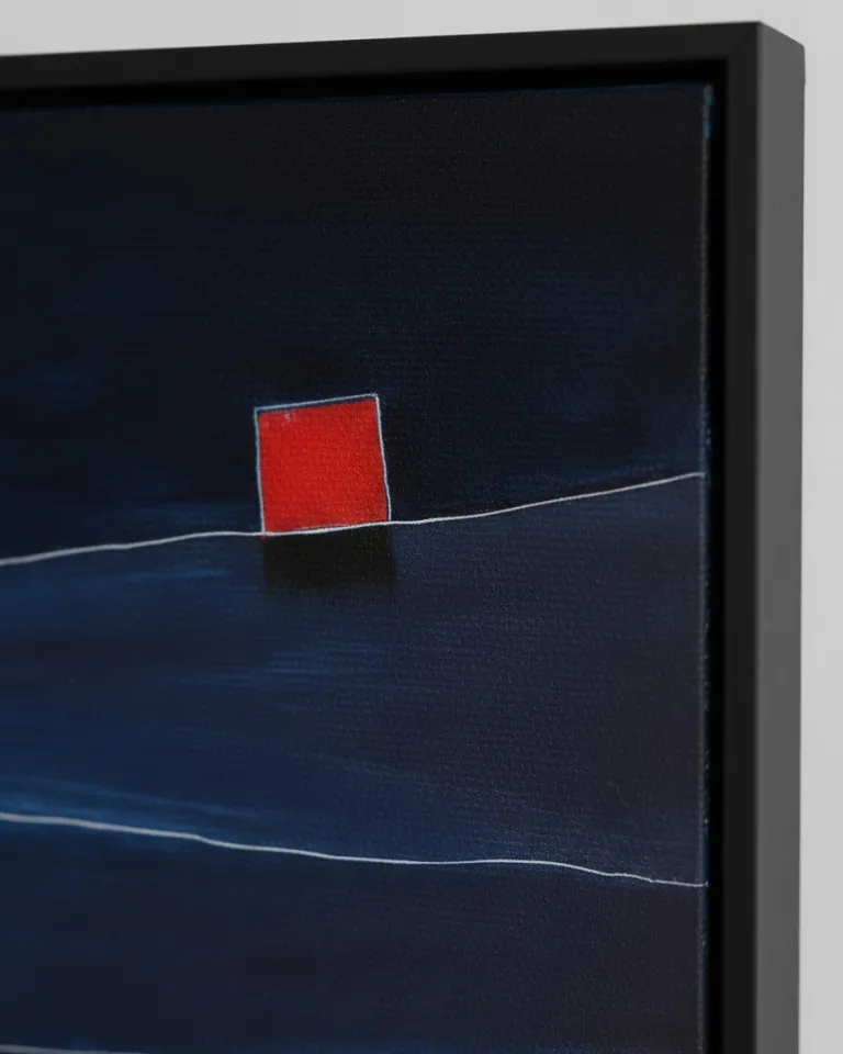 Close-up of an abstract painting in a black floating frame against a white wall. The texture of the dark blue and black painted canvas is visible, crossed horizontally by a thin white line upon which sits a small, bright red square with a dark shadow beneath it.