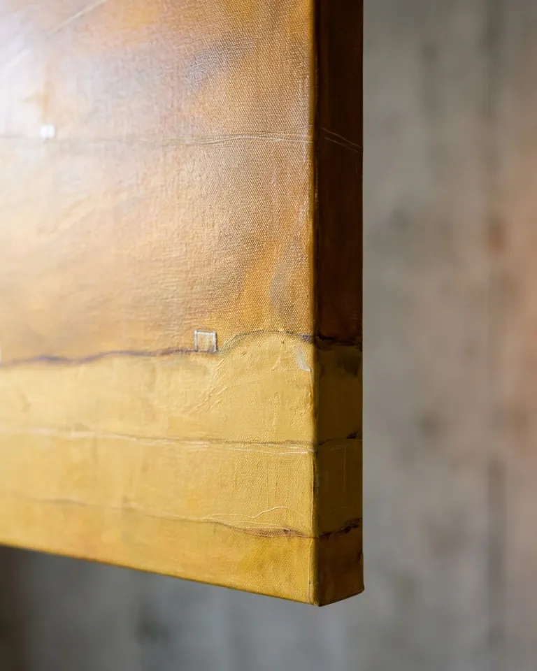 Close-up view of the edge of the abstract painting 'STRATA V — QUINQUE'. Visible acrylic and oil textures in ochre and yellow tones extend from the canvas surface to its side. A rough gray concrete wall forms the background. Directional lighting highlights the impasto, incised lines, and small blocks, emphasizing the artwork's materiality.