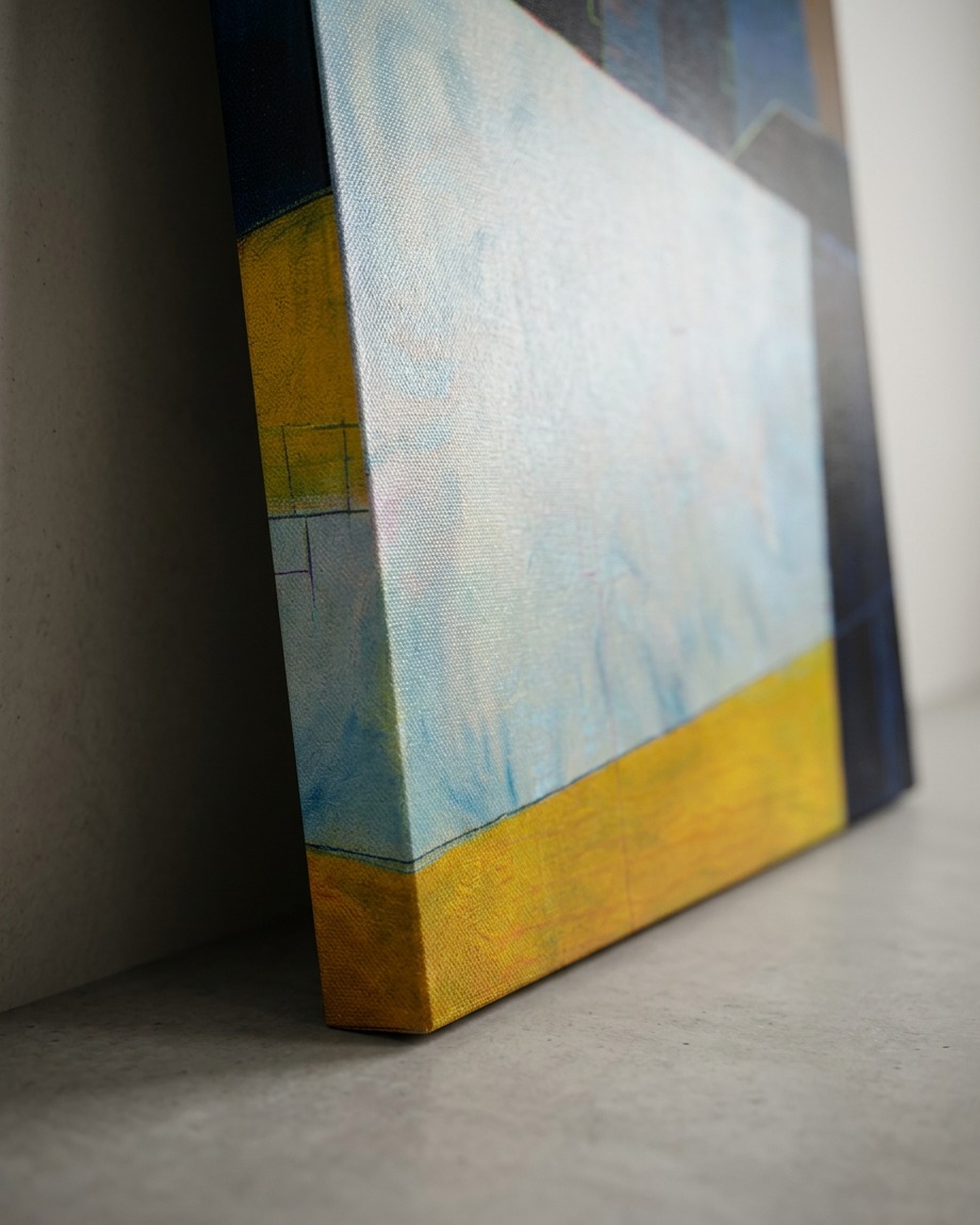 Close-up of a stretched canvas artwork with abstract geometric shapes in light blue, yellow-orange, and dark blue, leaning against a plain white wall on a gray concrete-like floor.