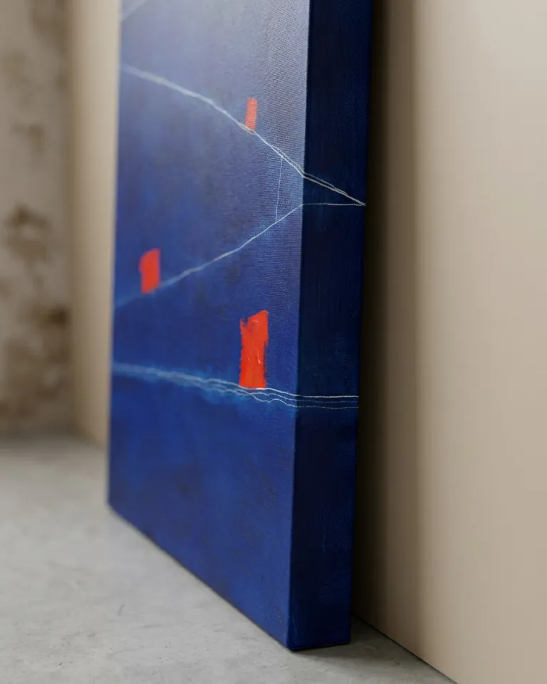 Close-up, slightly oblique view of Maurizio Valch's abstract painting "STRATA XII — SUBMERSA". A broad field of deep indigo blue covers most of the canvas. Fine, scraped white lines cross the surface in various directions. Three vibrant red rectangular blocks stand out against the blue background. The texture of the canvas and paint layers are visible, along with the continuation of the dark blue color on the artwork's side edge.