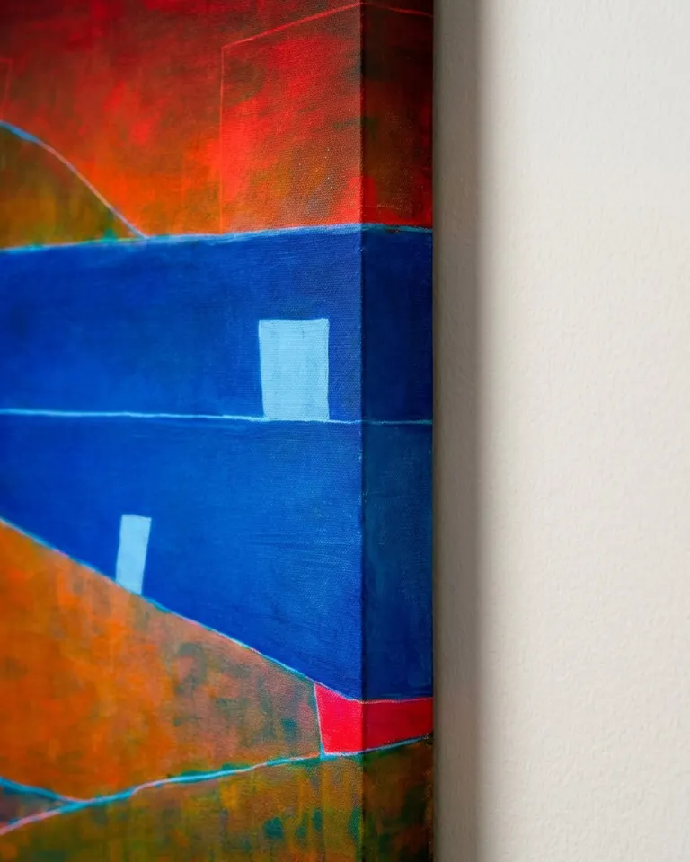 Detalle en primer plano lateral del borde de un cuadro de ochenta por ciento treinta centímetros. La iluminación nítida desde la derecha revela el espesor de tres centímetros del bastidor, donde la pintura de color azul se pliega continuamente hacia el lateral. En la cara frontal, se observan campos de color azul, ocre y un fragmento rojo carmesí superior con incisiones finas, proyectando una sombra sutil sobre la pared blanca.