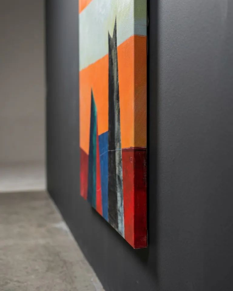 A close-up of the right edge of a vertical abstract painting. It shows a vibrant orange field, dark vertical forms, and a red block extending onto the side of the canvas. The artwork is mounted on a dark grey wall.