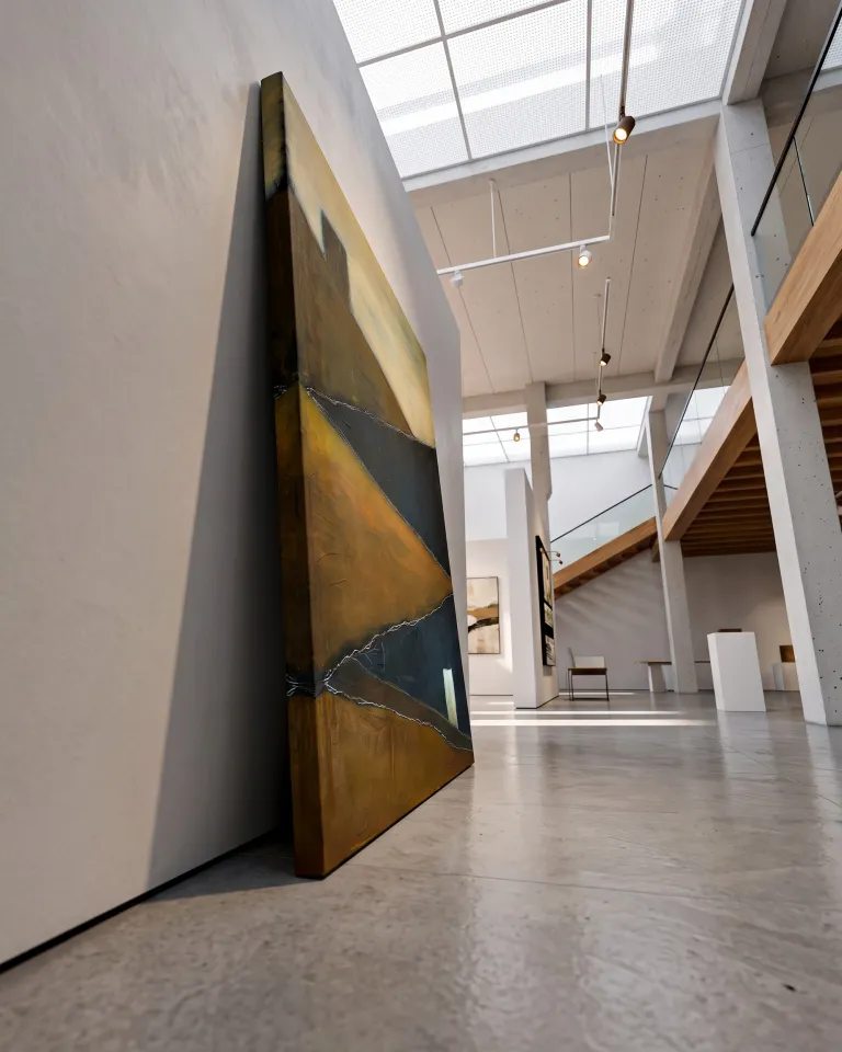 A low-angle photograph of a vertical abstract painting leaning directly against a gallery wall, resting on the floor. The artwork features diagonal planes in ochre and dark blue, partitioned by thin incised lines. In the background, a spacious contemporary art gallery is visible, with wooden beams, stairs, and a skylight casting soft, diffused light across the concrete interior.