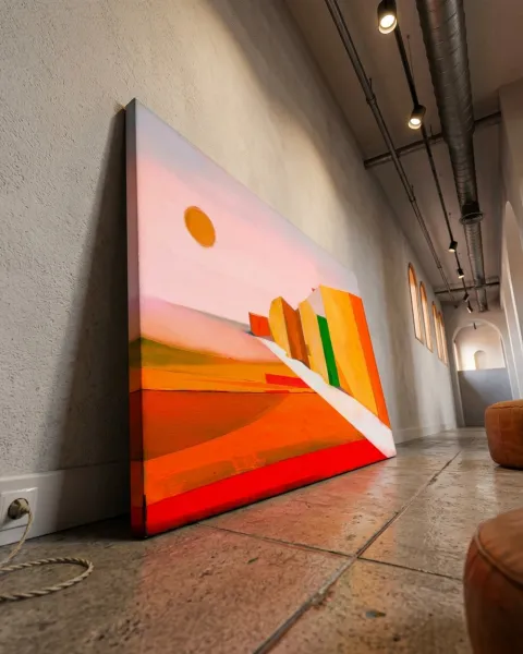 A horizontal abstract painting featuring angular, geometric structures in orange and yellow, receding into the background to create an architectural landscape. A white diagonal path crosses the composition towards the right, and a yellow circle appears in the upper left against a pale pink sky. Details such as a bright green rectangle and small, ladder-like incisions are visible on the surfaces of the structures.