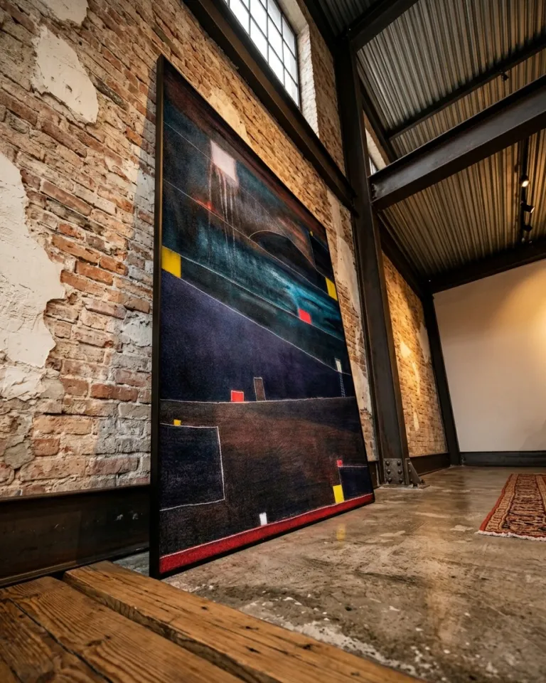 Vertical abstract painting 'ADITUS' by Maurizio Valch, leaning against an exposed brick wall. The artwork features dark blue and brown tones, with color fields, incised lines, and small red and yellow rectangles. A concrete floor and exposed steel beams of an industrial loft frame the scene under diffuse light.