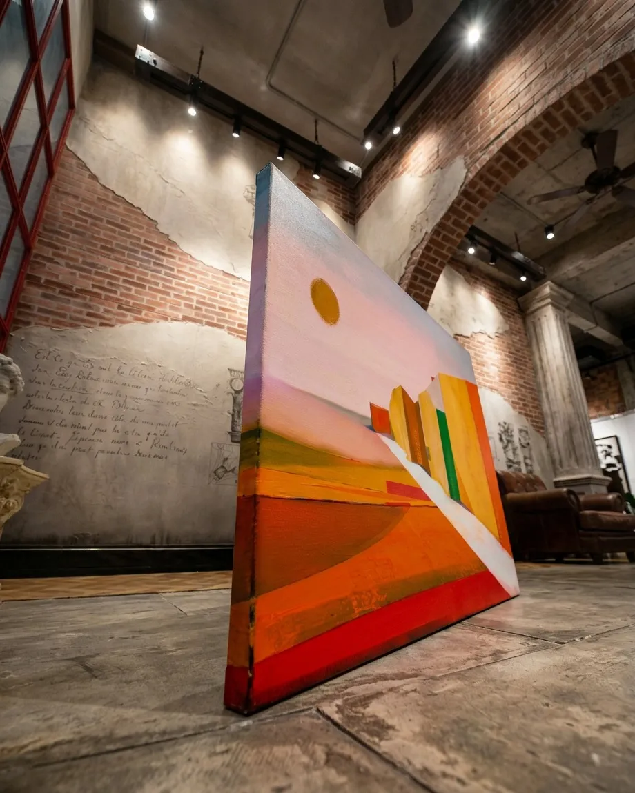 Horizontal abstract painting primarily in orange, yellow, and red. It features angular geometric structures receding along a diagonal white path. A yellow circle appears in the upper left against a pale sky. The surface shows visible texture and brushstrokes.