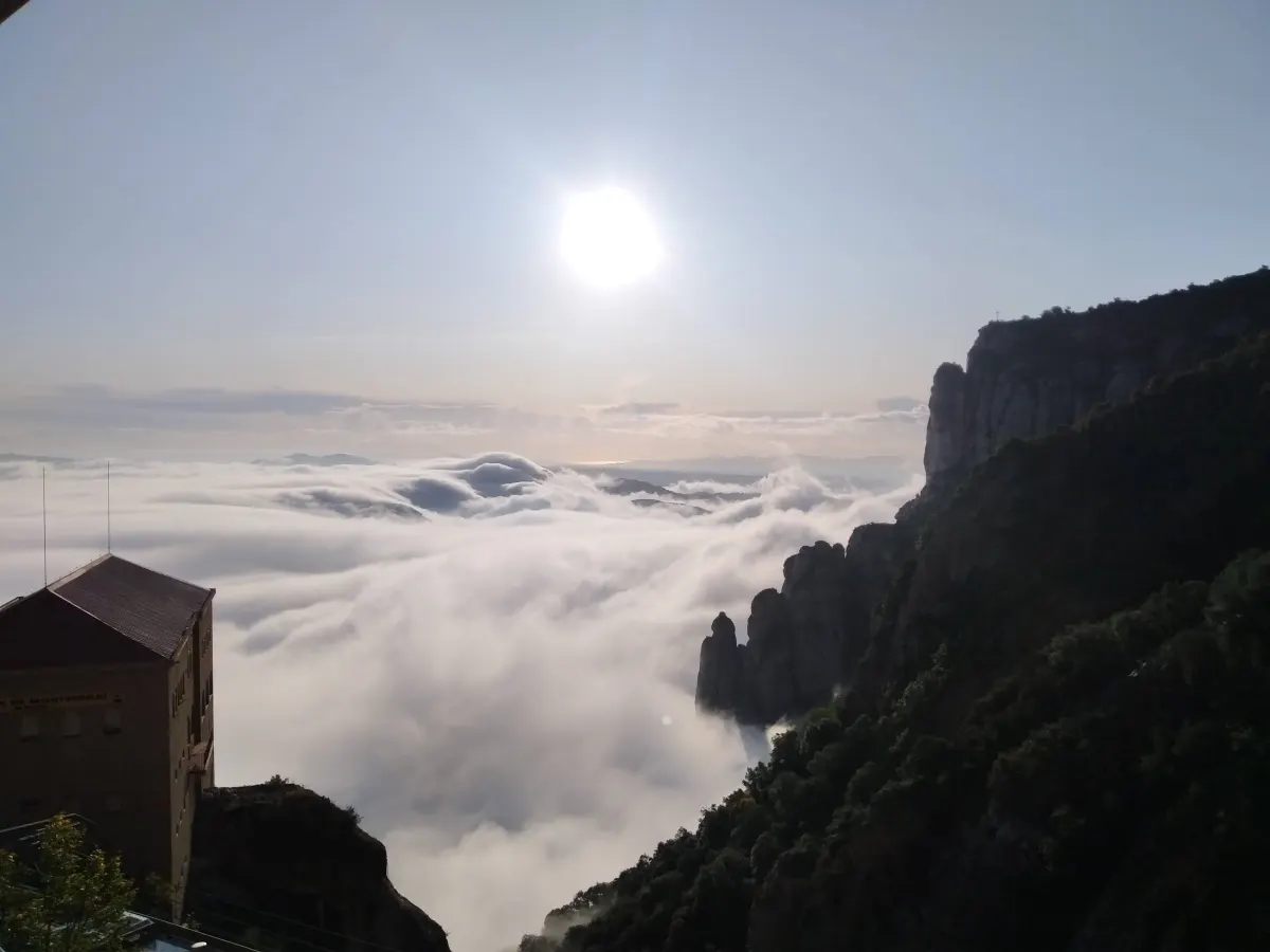 Aerial view of Montserrat mountain, Spain, with rocky peaks and a building on the left. A sea of white clouds covers the valley, while the sun shines brightly in the clear sky, illuminating the landscape.